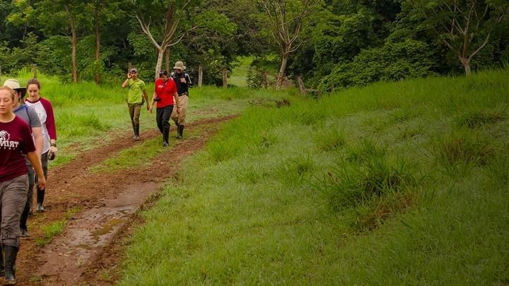 students hike along a muddy path in a lush field